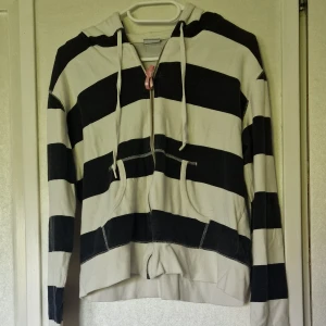 Randig hoodie från Gina Tricot - Svartvit randig hoodie från Gina Tricot i storlek M. Tröjan har dragkedja framtill, huva med snörning och två fickor. Tillverkad i bomull, perfekt för chill dagar. Tröjan är blekt i färgen, nopprig och har tappat spänsten i nederkanten. 