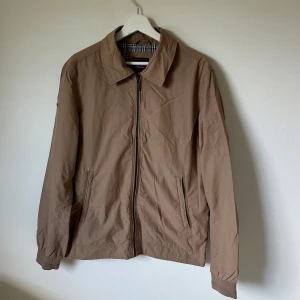 Beige vindjacka från Superdry - Snygg beige vindjacka från Superdry med klassisk krage och dragkedja framtill. Jackan har två fickor med dragkedja och rutigt innerfoder i blått, vitt och rött. Tillverkad i nylon och bomull, perfekt för vår och höst. Långärmad och stilren design. 2H4