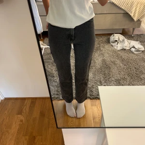 Svarta raka jeans med hög midja - Säljer ett par svarta jeans med rak passform och hög midja. Jeansen har klassisk femficksdesign, bälteshällor och knappgylf. Materialet är denim i bomull och färgen är tvättad svart för en cool look.
