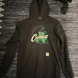 Svart Carhartt hoodie med tryck - Svart hoodie från Carhartt med stort grönt och gult tryck framtill, inklusive fyrklöver och logga. Klassisk känguruficka och huva med snörhål. Perfekt för dig som gillar streetwear och vill ha en skön och stilren tröja.
