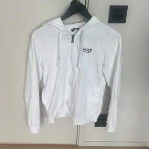 Vit hoodie från EA7 Emporio Armani - Snygg vit hoodie från EA7 Emporio Armani med dragkedja framtill, huva med snörning och diskret logga på bröstet. Tillverkad i mjuk bomull och har långa ärmar samt fickor på sidorna. Perfekt för en clean och sportig look. Priset är diskuterbart