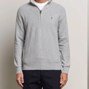 Grå half zip tröja från Polo Ralph Lauren - Snygg grå långärmad tröja från Polo Ralph Lauren med half zip och broderad logga på bröstet. Tröjan är i mjuk bomull och har ribbade muddar vid ärmslut och nederkant. Perfekt för lager-på-lager och stilren vardagslook.
