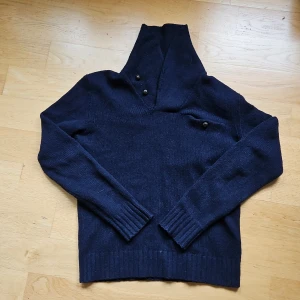 Mörkblå stickad tröja Polo Ralph Lauren - Mörkblå stickad tröja från Polo Ralph Lauren med hög krage och knappar vid halsen. Tröjan har lång ärm, ribbade muddar och en liten bröstficka med knapp. Perfekt för kyliga dagar och snygg till jeans.