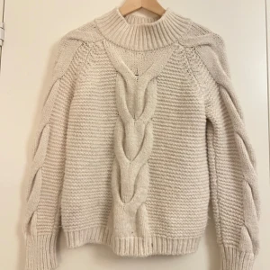 Beige stickad polotröja med flätmönster - Supermysig beige stickad polotröja med grovt flätmönster både framtill och längs ärmarna. Tröjan har en hög, ribbad krage och långa ärmar med breda muddar. Perfekt för kalla dagar när du vill vara både varm och snygg.