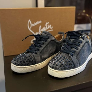 Christian Louboutin sneakers med nitar - Mörkblå sneakers från Christian Louboutin i mocka med svarta nitar på tån och klassisk röd sula. Skorna har snörning och vit platt sula, vilket ger en edgy och lyxig vibe. Perfekta för dig som vill sticka ut med exklusiva detaljer och streetstyle-känsla.