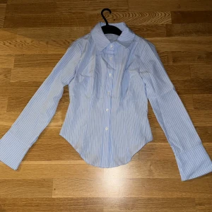 Blåvit randig skjorta med vida ärmar - Snygg blå skjorta med vita ränder och klassisk krage. Skjortan har långa, vida ärmar och knäppning framtill med vita knappar. Figursydd passform och lätt rundad nederkant. Perfekt för en clean och stilren look.