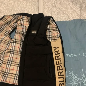 Svart dunväst från Burberry, M - Säljer en svart dunväst från Burberry med klassiskt rutigt foder i beige, rött och svart. Västen har en stor beige panel med Burberry-logga längs sidan och dragkedja framtill. Perfekt för lager-på-lager och riktigt snygg streetstil.