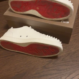 Christian Louboutin vita sneakers med nitar - Säljer ett par vita sneakers från Christian Louboutin med ikoniska röda sulor och coola nitar på tån. Skorna är i skinn och har snörning. Perfekta för dig som vill sticka ut med en lyxig och grischig stil.