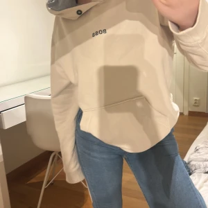 Beige hoodie från BOSS med huva - Säljer en beige hoodie från BOSS med svart broderad logga på bröstet. Tröjan har en stor huva med grå insida, känguruficka framtill och långa ärmar. Perfekt för en chill och avslappnad stil.
