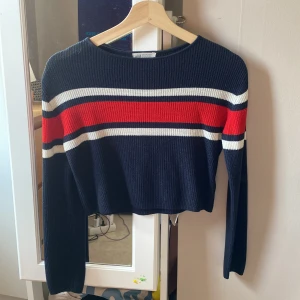 Randig croppad tröja från H&M - Säljer en ribbad, croppad tröja från H&M med marinblå bas och breda ränder i rött, vitt och blått över bröstet. Tröjan har lång ärm och rund halsringning, perfekt för dig som gillar sportig och trendig stil.