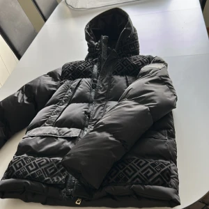 Svart pufferjacka från Glorious Gangsta - Säljer en svart pufferjacka från Glorious Gangsta med feta mönstrade detaljer på axlar, huva och nederkant. Jackan har huva, dragkedja och knappar framtill samt stora fickor. Materialet är polyester och jackan har en riktigt clean och modern vibe.