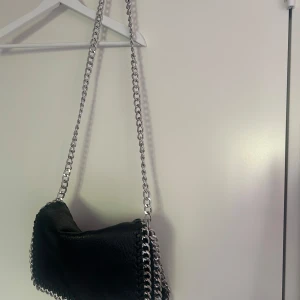 Svart axelväska med kedja från Tiamo - Snygg svart axelväska från Tiamo i skinnimitation med grov silverfärgad kedja som axelrem och detaljer längs kanterna. Väskan har dragkedja upptill och en stilren, modern look som passar till många outfits.