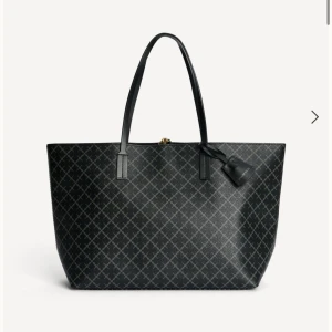 Svart shoppingväska från By Malene Birger - Stilren svart shoppingväska från By Malene Birger med klassiskt rutigt mönster och långa handtag. Väskan är rymlig och har detaljer i guld samt en tag med logga. Tillverkad i slitstarkt syntetmaterial med elegant finish.