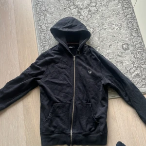 Svart hoodie från Fred Perry med dragkedja - Svart hoodie från Fred Perry med klassisk logga broderad på bröstet. Tröjan har huva, hel dragkedja framtill och två fickor. Tillverkad i mjuk bomullsmix och har ribbade muddar. Perfekt för en chill och stilren look.