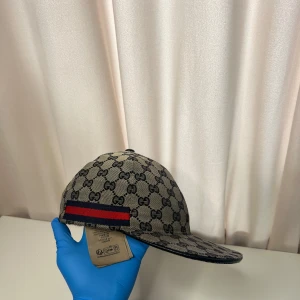 Gucci keps med GG-mönster och röd-blå detalj - Säljer en beige keps från Gucci med klassiskt GG-monogram över hela ytan. Kepsen har en röd och marinblå rand på sidan och justerbart spänne bak i svart skinn. Materialet är bomull och kepsen har en böjd skärm. Perfekt för dig som gillar lyxiga accessoarer.