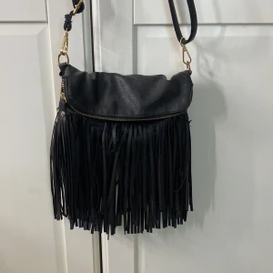 Svart axelväska med fransar - Cool svart axelväska i skinnimitation med långa fransar framtill och dragkedja upptill. Väskan har justerbar axelrem med guldfärgade metalldetaljer. Perfekt för dig som vill ha en trendig och unik accessoar till din outfit.