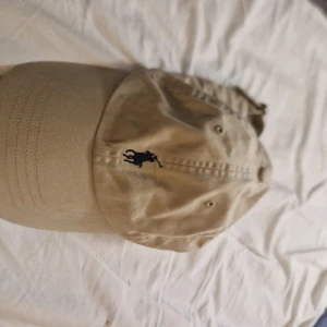 Beige keps från Polo Ralph Lauren - Snygg beige keps från Polo Ralph Lauren med klassisk broderad logga i svart framtill. Kepsen har böjd skärm och justerbar rem baktill för perfekt passform. Tillverkad i mjuk bomull som är skön att bära hela dagen.