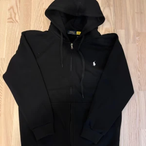 Svart Polo Ralph Lauren zipper - Svart hoodie från Polo Ralph Lauren med dragkedja och huva. Klassisk broderad logga i vitt på bröstet och snörning i huvan. Mjuk bomullskänsla och ribbade muddar vid ärmslut och nederkant. Perfekt för en chill och stilren look.