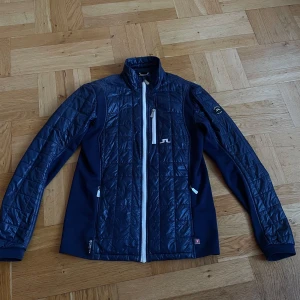 Mörkblå quiltad jacka från J.Lindeberg - Snygg mörkblå jacka från J.Lindeberg med quiltade partier framtill och på ärmarna. Jackan har hel dragkedja i vitt, ståkrage och flera praktiska fickor. Materialet är polyester och jackan har en sportig look som passar perfekt till kyligare dagar.