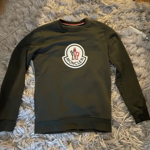 Mörkgrön sweatshirt från Moncler - Säljer en mörkgrön sweatshirt från Moncler med stort vitt och rött logotryck på bröstet och mindre logga på ärmen. Tröjan har rund halsringning och långa ärmar. Perfekt för dig som gillar stilrena och exklusiva plagg. Tröjan är använd fåtal gångånger är i bra skick inga defekter 
