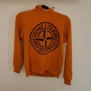 Orange hoodie från Stone Island Junior - Snygg orange hoodie från Stone Island Junior med stort svart broderat logotyptryck på bröstet. Tröjan har huva, långa ärmar och är tillverkad i mjuk bomull. Perfekt för dig som vill sticka ut med en färgstark och trendig look. Den är bäst när man går på fotboll!! 