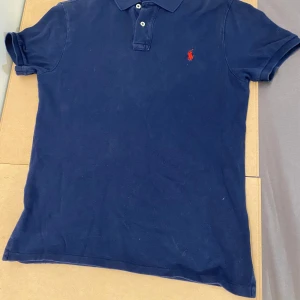 Marinblå pikétröja från Ralph Lauren - Snygg marinblå pikétröja från Polo Ralph Lauren i slim fit-modell. Klassisk design med krage, knappar och röd broderad logga på bröstet. Den är M i barn storlek så den passar personer som är ca 163-170