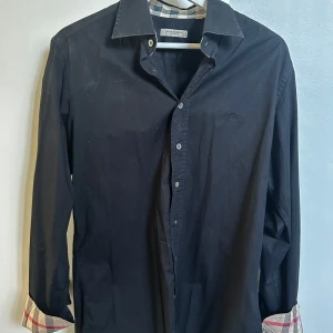 Svart Burberry skjorta med rutig detalj - Svart skjorta från Burberry i bomull med klassisk rutig insida på krage och manschetter i beige, rött och grått. Broderad Burberry-logga på bröstet, knappar framtill och långärmad modell. Perfekt för dig som gillar stilrena plagg med lyxig touch.