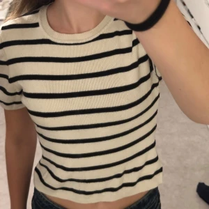 Randig t-shirt från Gina Tricot - Superfin kortärmad t-shirt från Gina Tricot i beige med svarta horisontella ränder. Modellen är croppad med rund halsringning och mjukt, stickat material som känns skönt mot huden. Perfekt till jeans eller kjol för en chill look.
