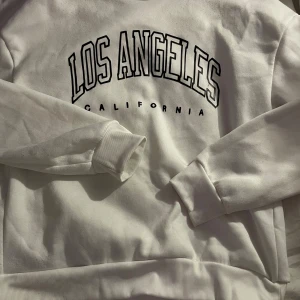Vit sweatshirt Los Angeles California - Vit sweatshirt med svart tryck 'Los Angeles California' på bröstet. Tröjan har rund hals, ribbade muddar vid ärmslut och nederkant samt lång ärm. Perfekt för en avslappnad och trendig look.