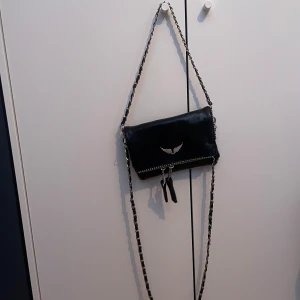 Svart Zadig & Voltaire axelväska - En svart Zadig liknande axelväska i skinn med silverfärgade kedjedetaljer och dragkedja. Väskan har en ikonisk vinge framtill och snygga nitar längs kanten. Perfekt storlek för det viktigaste och med ett foder med logga inuti.