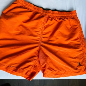 Orange badshorts från Polo Ralph Lauren - Snygga orange badshorts från Polo Ralph Lauren med elastisk midja i grönt och innerbyxa i mesh. Klassisk design med bakficka och broderad logga framtill. Perfekta för stranden eller poolen.