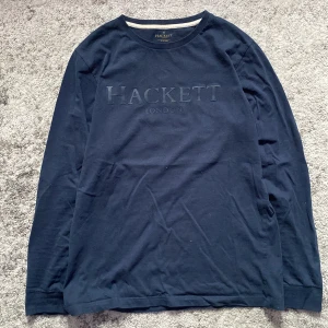 Mörkblå långärmad tröja Hackett - Snygg mörkblå långärmad tröja från Hackett London med diskret logga framtill. Tröjan har rund halsringning med vit detalj på insidan och är gjord i mjuk bomull. Perfekt för dig som gillar stilrena och klassiska plagg.