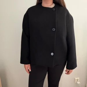 Svart ulljacka från BikBok, storlek L - Stilren svart jacka från BikBok i ullblandning. Jackan har rak, boxig passform, knäpps med tre stora knappar framtill och har rund halsringning. Perfekt för dig som gillar minimalistisk och clean stil. Aldrig använd, pris kan diskuteras!