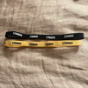 Stronger diadem svart och gul - Två elastiska diadem från Stronger, ett svart och ett gult, båda med vit text 'STRONGER' tryckt runt om. Perfekta för att hålla håret på plats under träning eller vardag. Mjukt och stretchigt material som sitter bekvämt på huvudet.