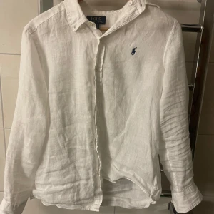 Vit linneskjorta från Polo Ralph Lauren - Klassisk vit skjorta i linne från Polo Ralph Lauren med blå broderad logga på bröstet. Skjortan har knappar framtill, button down-krage och långa ärmar. Perfekt för en clean och stilren look.