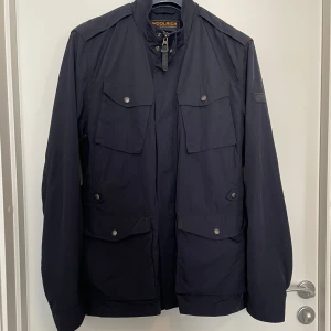 Svart vindjacka från Woolrich XL - Snygg svart vindjacka från Woolrich med fyra stora fickor framtill och hög krage. Jackan har diskret logotyp på ärmen och stängs med dragkedja och tryckknappar. Perfekt för dig som gillar stilren och funktionell design. Pris kan diskuteras!