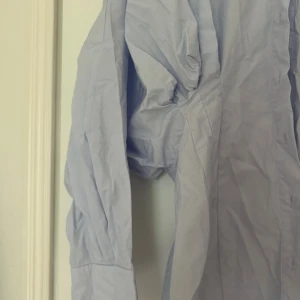 Ljusblå oversized skjorta/klänning - Säljer en ljusblå oversized skjorta med klassisk krage och knäppning framtill. Skjortan har långa ärmar med manschett och är tillverkad i ett tunt bomullstyg. Perfekt för en avslappnad och trendig look.