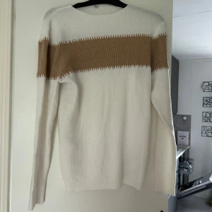 Vit och beige ribbstickad tröja med kjol  - Ribbstickad tröja i vitt med en bred beige rand över bröstet och ärmarna. Tröjan har rund halsringning och långa ärmar. Perfekt för dig som gillar en enkel men snygg stil med lite retrovibbar.