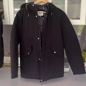 Svart parkajacka Jack & Jones S - Svart parkajacka från Jack & Jones med huva, dragkedja och tryckknappar. Jackan har två stora fickor framtill med lock och snygga detaljer i orange vid snörningen. Fodrad insida för extra värme, perfekt för kyliga dagar.