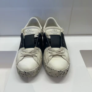 Valentino Garavani Open sneakers 37,5 - Snygga Valentino Garavani Open sneakers i vitt skinn med svart band över vristen och cool stänkmönstrad sula. Klassisk logga på plösen och texten 'OPEN FOR A CHANGE' på sidan. Skorna har snörning och rund tå för en clean look.