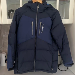 Mörkblå pufferjacka från Sail Racing - Säljer en mörkblå pufferjacka från Sail Racing med huva och två vertikala bröstfickor med dragkedja. Jackan har en sportig look, dubbla dragkedjor framtill och logga på bröstet. Perfekt för kalla dagar och riktigt snygg till streetwear.