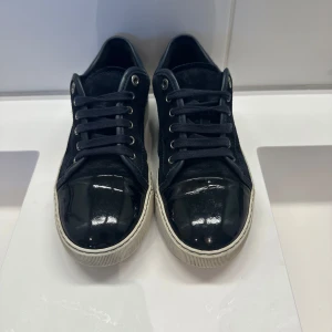 Svarta sneakers från Lanvin i mocka - Snygga svarta sneakers från Lanvin med ovandel i mocka och lackad tå. Skorna har vita sulor, svarta skosnören och metallfärgade öljetter. Perfekta för dig som gillar stilrena och exklusiva sneakers med en twist. Storlek UK6. Box fås med!!