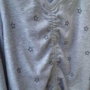 Grå långärmad tröja med stjärnor - Grå ribbad långärmad tröja med svarta stjärnor över hela tyget. Tröjan har v-ringning och snygg snörning framtill som ger en rynkad effekt. Perfekt för dig som gillar en chill och trendig stil.