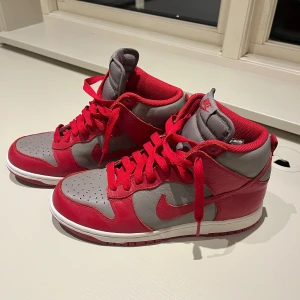 Nike Dunk High röda UNLV - Nike Dunk High sneakers i rött och grått med klassisk vit sula. Skorna har högt skaft, snörning och är tillverkade i skinn. Röd Nike swoosh på sidan och röd yttersula. Perfekta för dig som gillar streetwear och vill sticka ut med färg.                     Nypris: 3500-5000kr