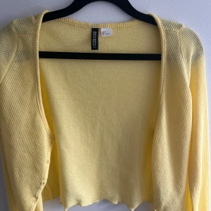 Gul stickad kofta  - Söt ljusgul stickad kofta från Divided med långärmad design och öppen framtill. Perfekt att slänga över en topp för en fräsch vårlook. Mjuk och lätt i materialet, med vågig nederkant som ger en extra touch.