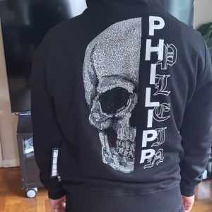 Svart Philipp Plein hoodie med dödskalle - Svart hoodie från Philipp Plein med dragkedja, huva och snörning. På ryggen finns en stor glittrig dödskalle och texten PHILIPP PLEIN i vitt. Liten logga på bröstet. Grym streetstyle med coola detaljer och bekväm passform. Använt den 3 gånger och köpte den från MIINTO.