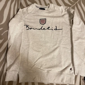 Ljusgrå sweatshirt från Bondelid - Ljusgrå sweatshirt från Bondelid med svart broderad logga och ett litet sköldmärke framtill. Tröjan har rund hals, långa ärmar och ribbade muddar vid ärmslut och nederkant. Perfekt för en chill och stilren look.