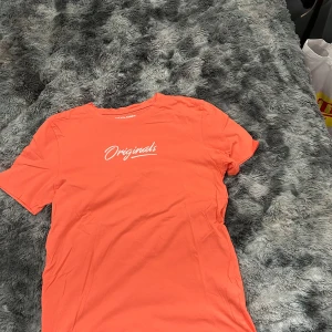 Orange t-shirt från Jack & Jones - Snygg orange t-shirt från Jack & Jones Originals med vit text på bröstet. Klassisk rund hals och korta ärmar. Tillverkad i mjuk bomull som känns skön mot huden. Perfekt för dig som gillar en enkel men stilren look.