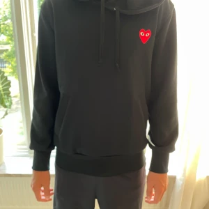 Svart Comme des Garçons Play hoodie - Svart hoodie från Comme des Garçons Play med det ikoniska röda hjärtat med ögon på bröstet. Tröjan har huva med snörning, magficka och ribbade muddar. Perfekt för dig som gillar streetwear och vill ha något stilrent men ändå med en twist.