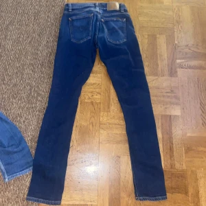 Mörkblå slim fit jeans från Nudie Jeans - Säljer ett par mörkblå slim fit jeans från Nudie Jeans i storlek 29/32. Klassisk femficksmodell med snygga kontrastsömmar och normalhög midja. Jeansen är tillverkade i stretchig bomull för en bekväm passform och har en stilren look.